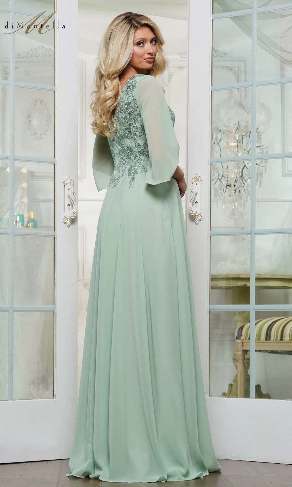 A woman with long blonde hair wears the Rina Di Montella RD3241, a light green chiffon formal dress with lace details and an elegant A-line skirt, standing in front of glass doors and looking over her shoulder.